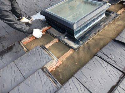 岐阜市K様邸の屋根修理施工風景。瓦職人が天窓（トップライト）周囲の既存瓦を手作業で一枚ずつ丁寧に剥がしている。露出した屋根下地（ルーフィング）や瓦を固定していた木材（桟木）が見える。